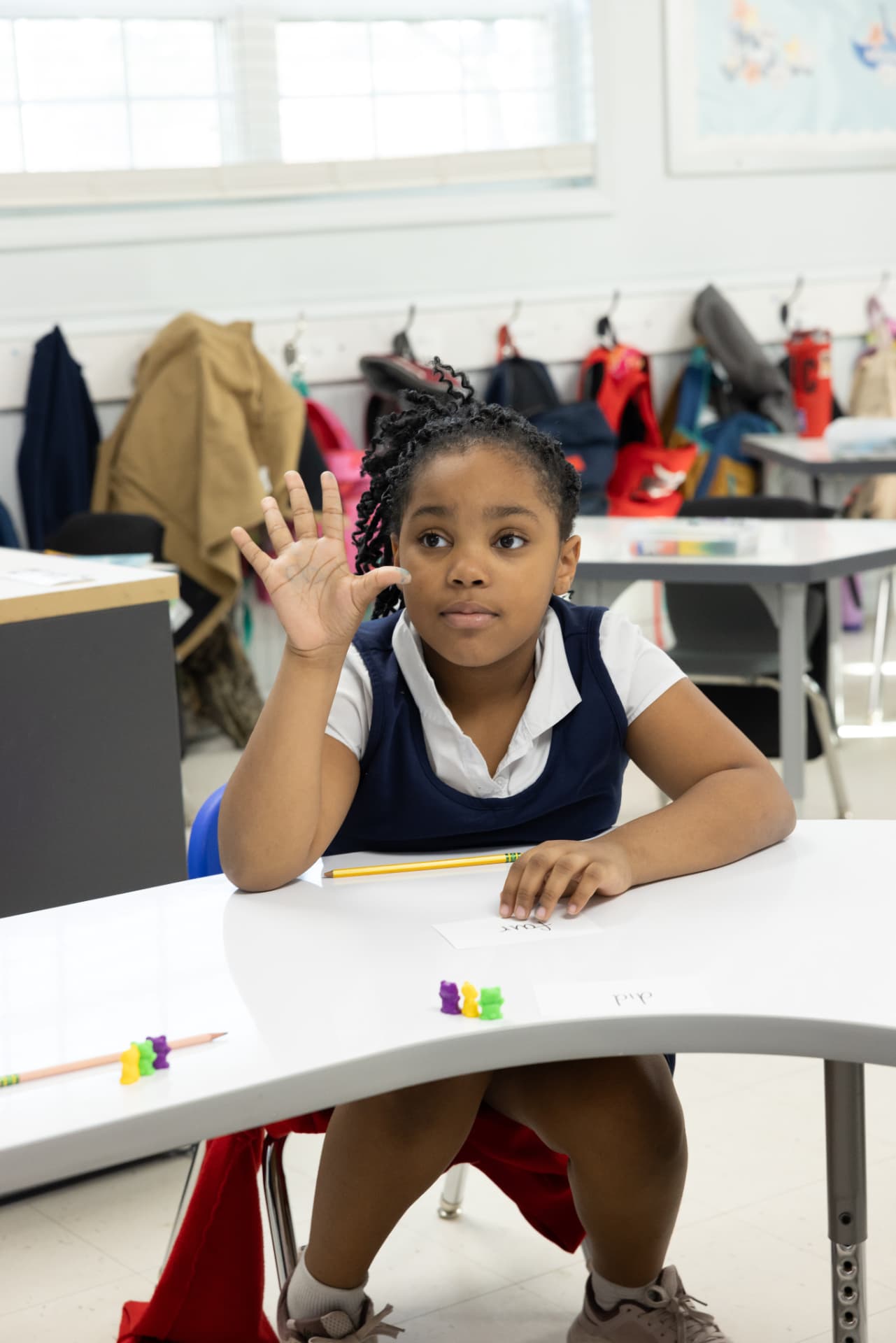A student raising her hand at her desk beside word cards and counting bears