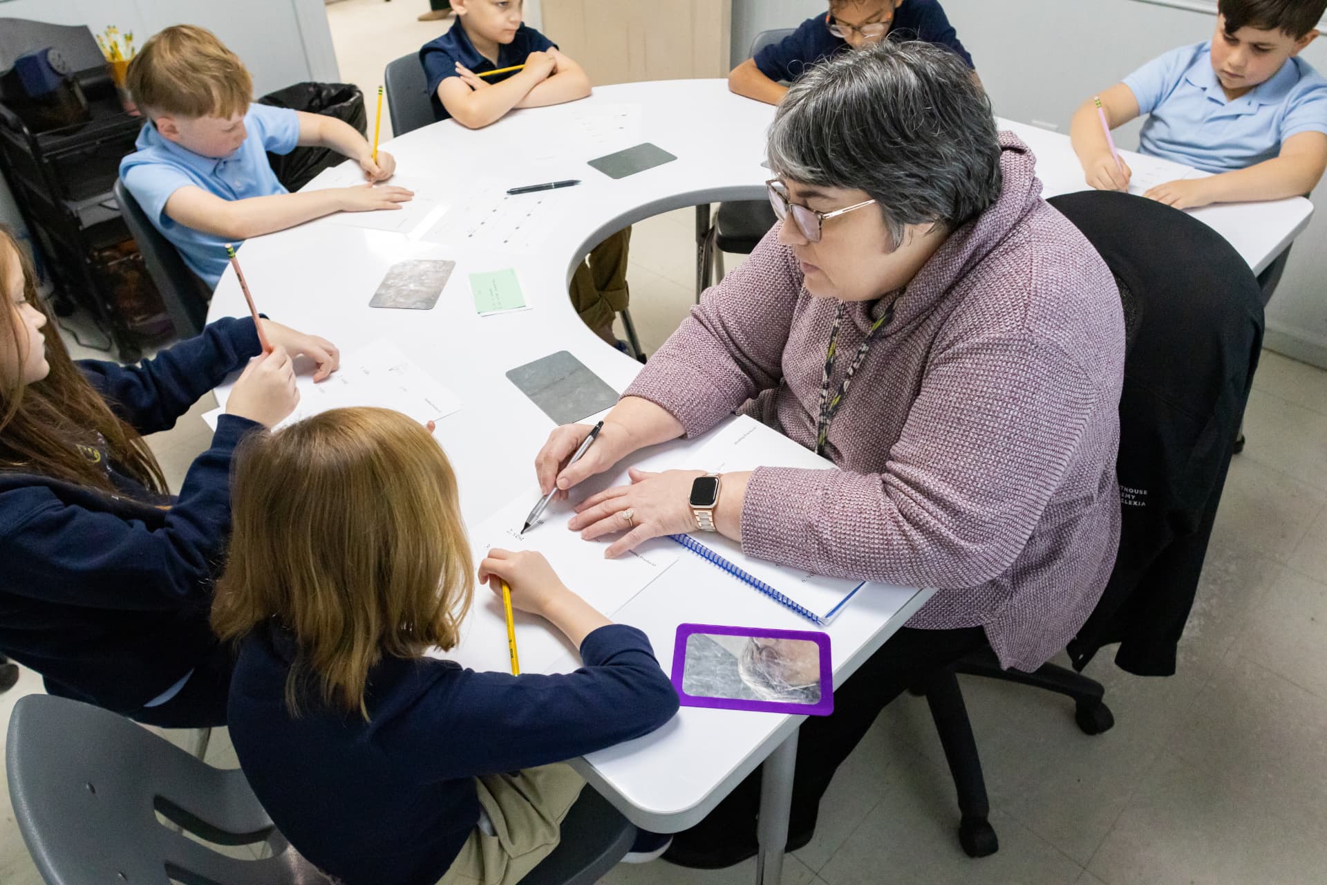 A teacher guiding six students through written work at a horseshoe table