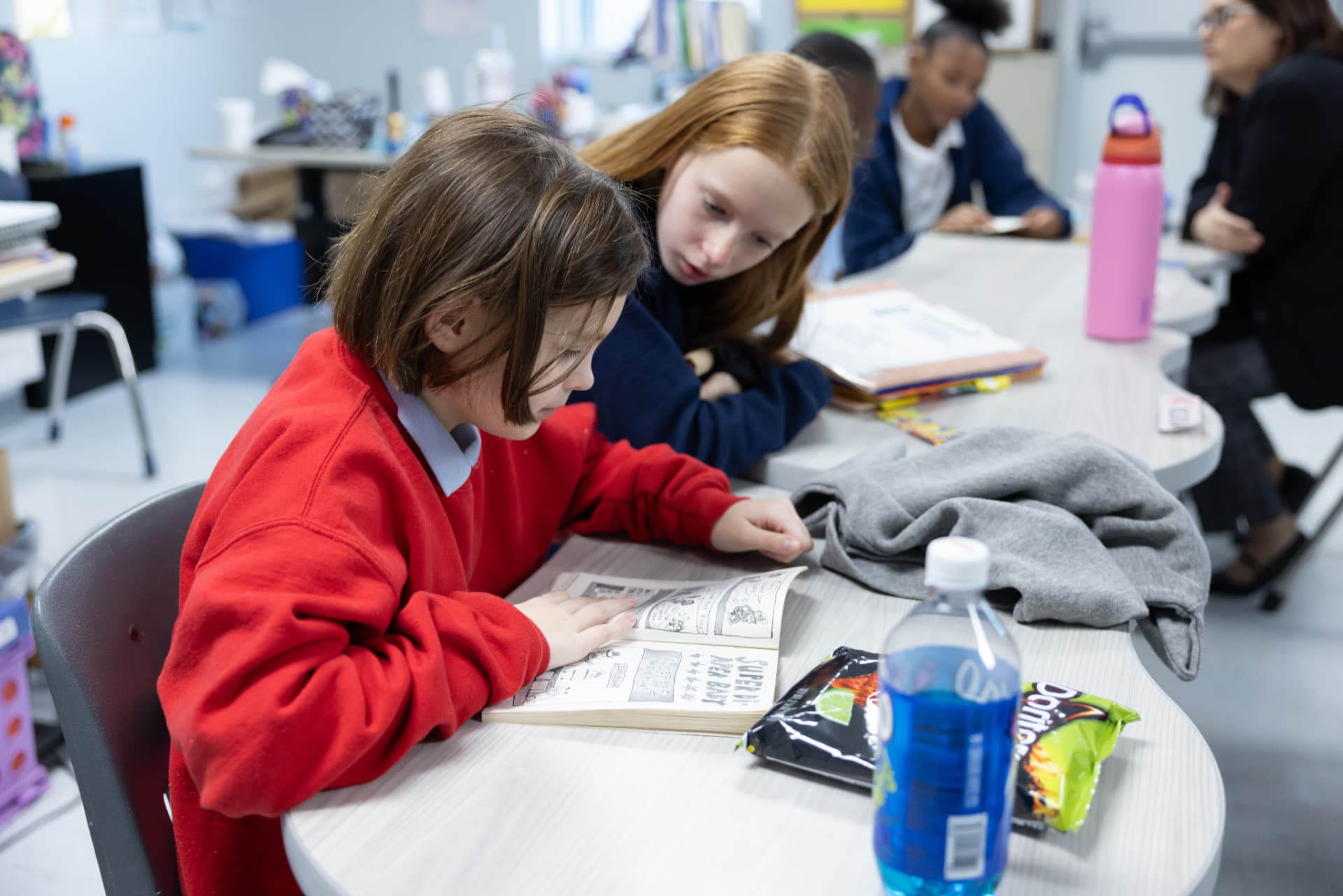 A student reading a graphic novel at a table while a classmate leans in to look