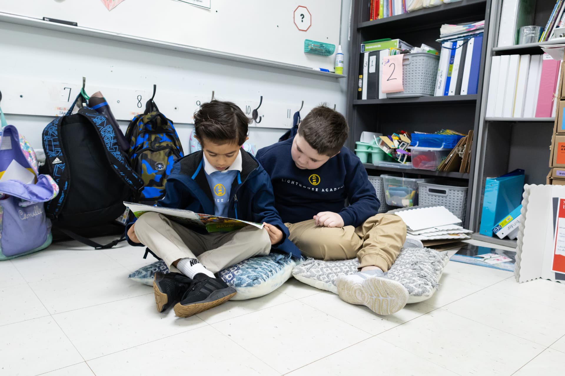 Two students reading side by side on floor cushions in a classroom reading corner