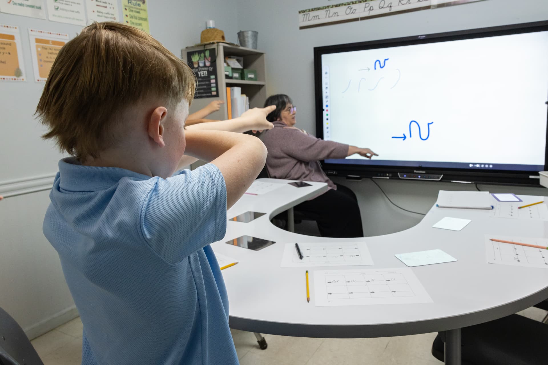 A student air-writing a cursive letter shape along with his teacher at the board
